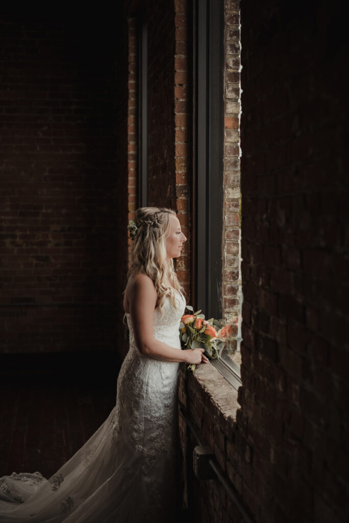 Wedding Photographer, A bride is in her wedding dress and looks outside the church building window as she leans on the brick window sill.